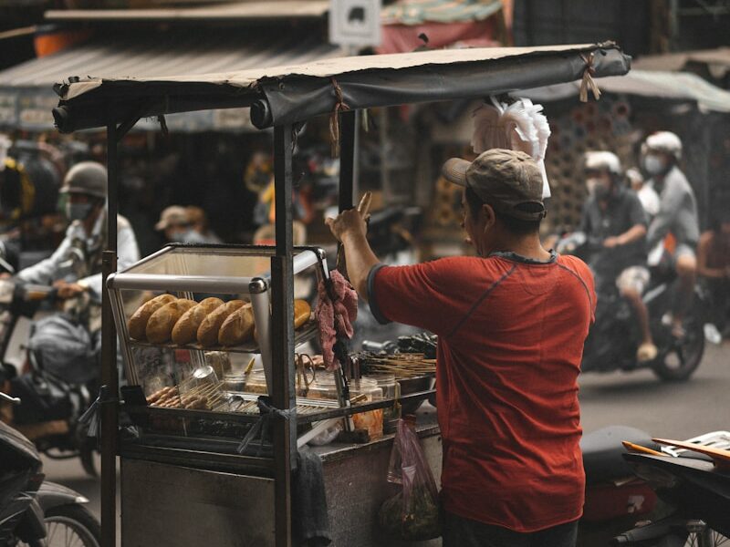 man in red t-shirt and black pants standing in front of food stall during daytime